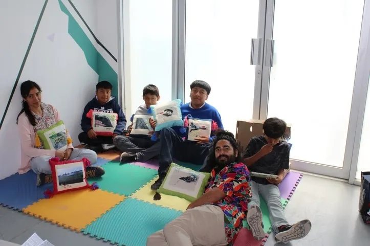 Niñas, niños y talleristas muestran sus cojines bordados sentados sobre un tapete de colores.